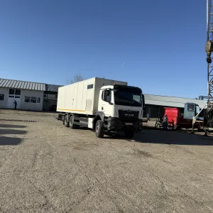 White box truck parked on a gravel lot near industrial buildings and drilling equipment.