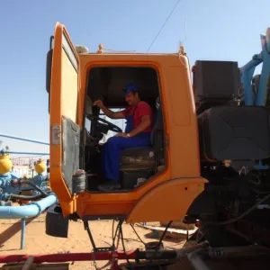 Worker in blue uniform and helmet operating machinery inside a yellow truck cab outdoors.